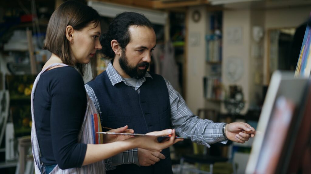 Artist guiding student painting at easel in studio.