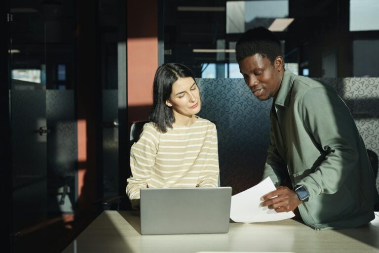 Two professionals discussing work over a laptop in a bright, modern office setting.