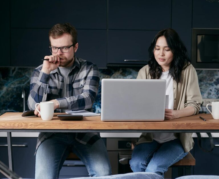 Couple working together in kitchen, focused on laptop tasks and discussions.